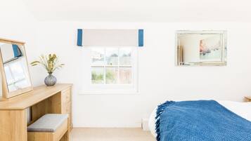 Une chambre avec un bureau en bois, une fenêtre avec des stores, un lit recouvert d’une couverture bleue et un tableau accroché au mur.