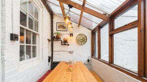 A wooden table with a bench, a potted plant, a clock, and a hanging light fixture in a room with white brick walls and large windows. - Shakespeare House, Free Parking, Town Centre Location (Stratford-upon-Avon)