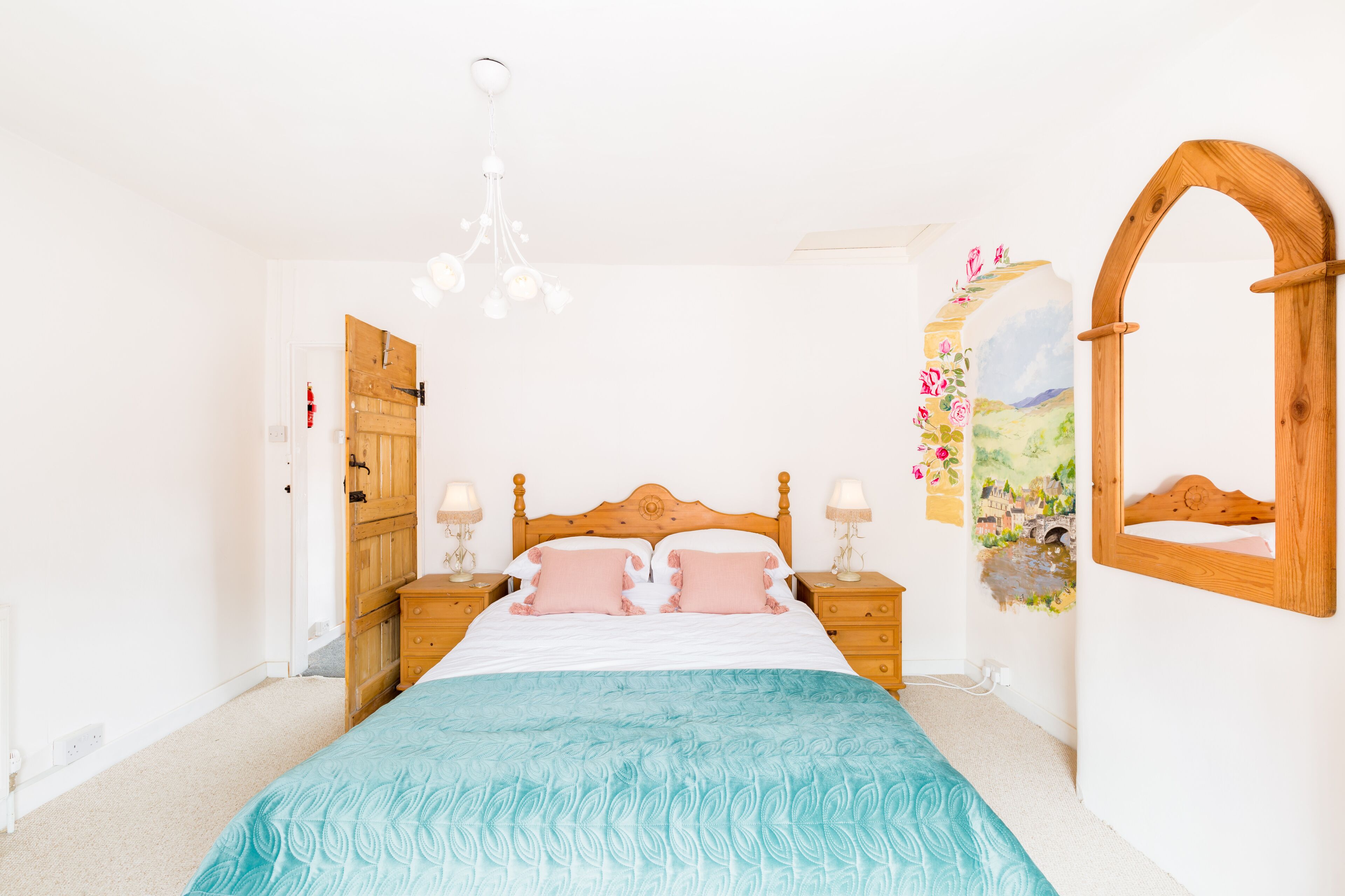 A bedroom with a wooden bed, two bedside tables with lamps, a wooden wardrobe, a wall-mounted mirror, and a floral artwork.