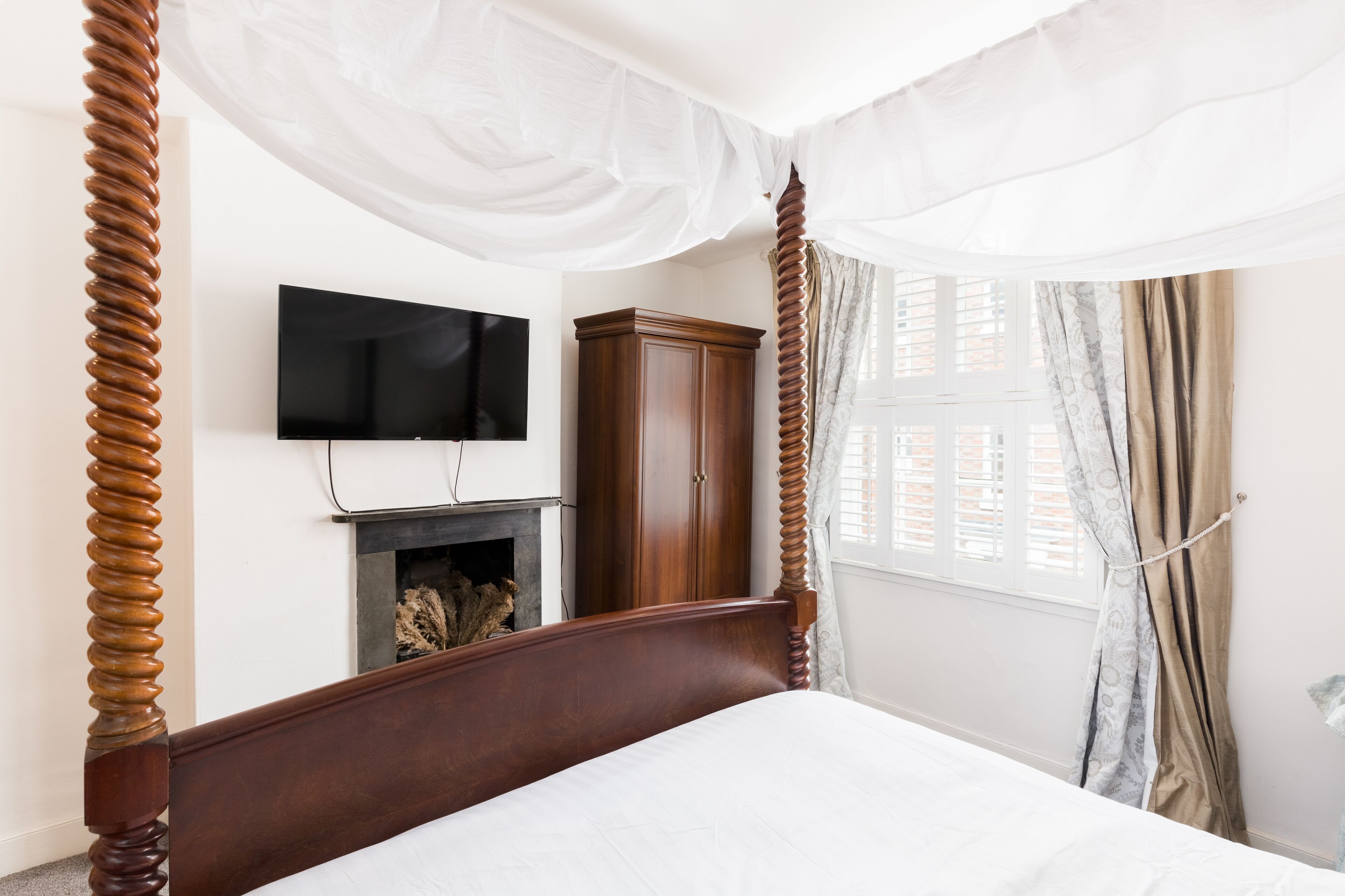 A bedroom with a four-poster bed, a television mounted on the wall, a wooden wardrobe, and a fireplace.