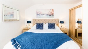 A neatly made bed with a blue blanket, two matching pillows, a headboard, and a painting above. There are two bedside tables with lamps and a wooden wardrobe with a mirror. - Shakespeare House, Free Parking, Town Centre Location (Stratford-upon-Avon)