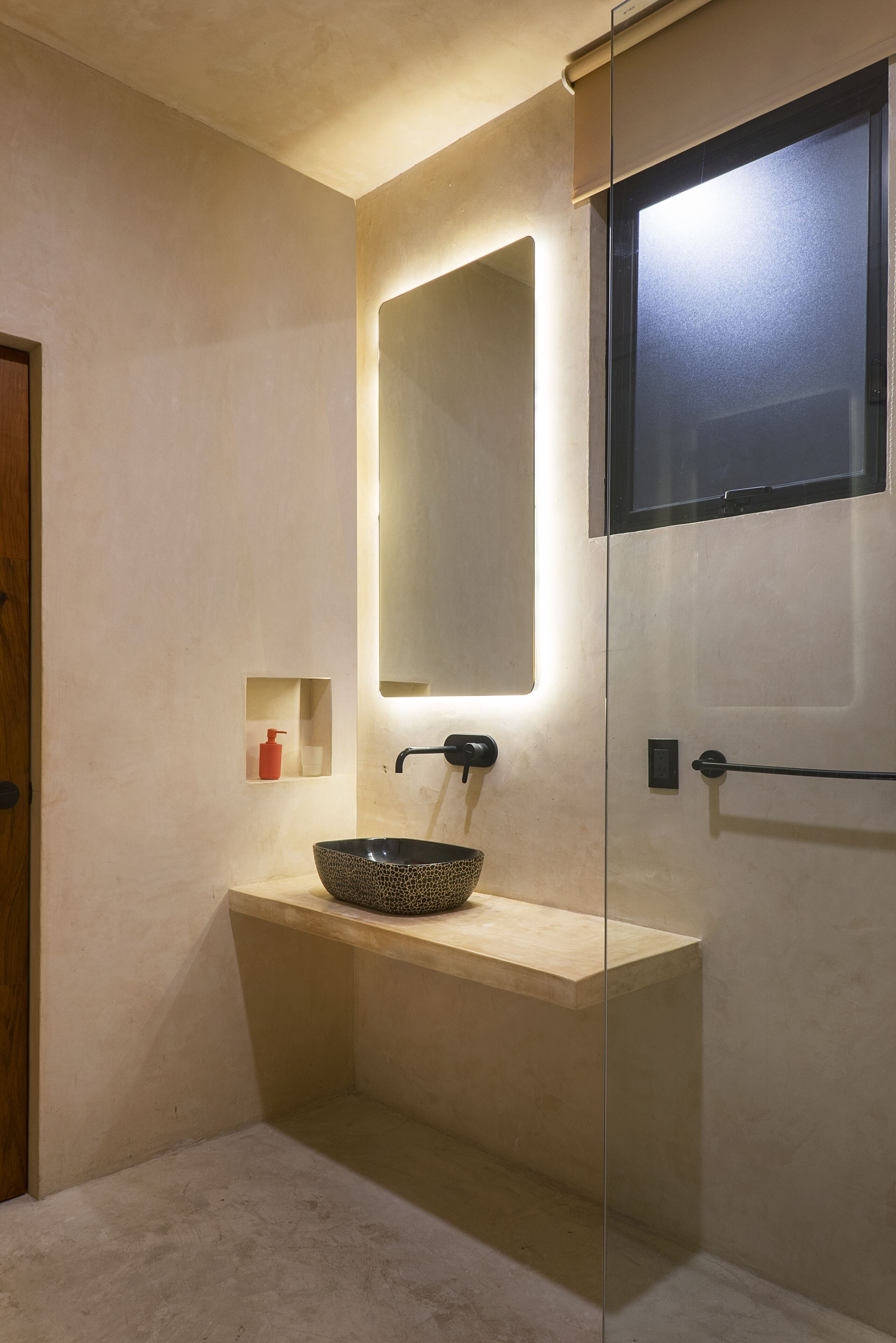A modern bathroom with a large mirror, a bowl sink, and a small red bottle on a shelf.