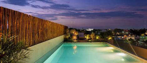 Piscina infinita en la azotea con un muro de madera y sillones de descanso, con vista a un paisaje urbano al anochecer.