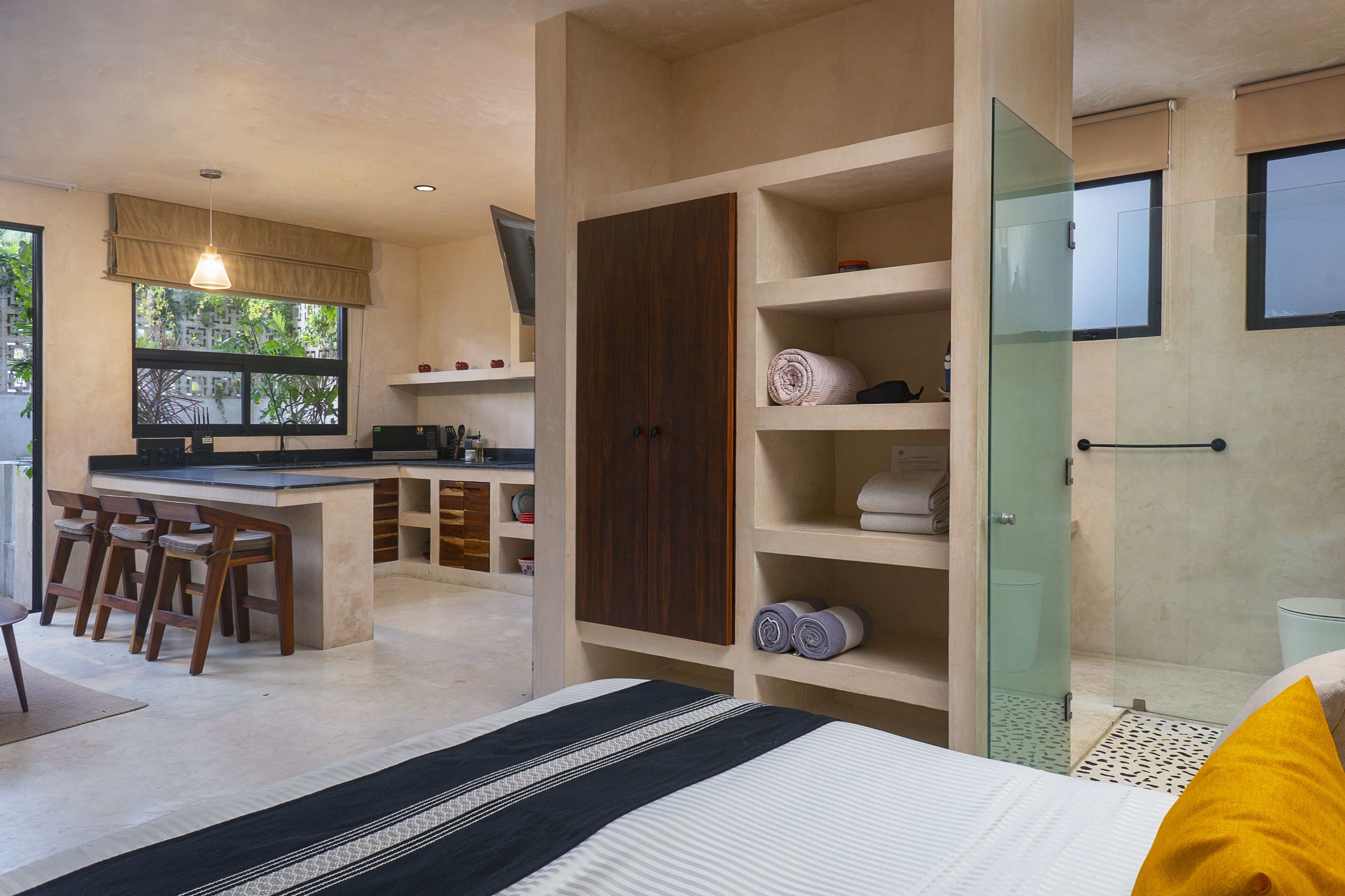 A modern hotel room with a large bed, a separate dining area with bar stools, a built-in wardrobe, and a shower area.