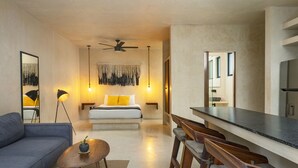 A modern hotel room with a sofa, a bed with yellow pillows, a ceiling fan, and a dining area with wooden benches. - UJO Tulum (Tulum)