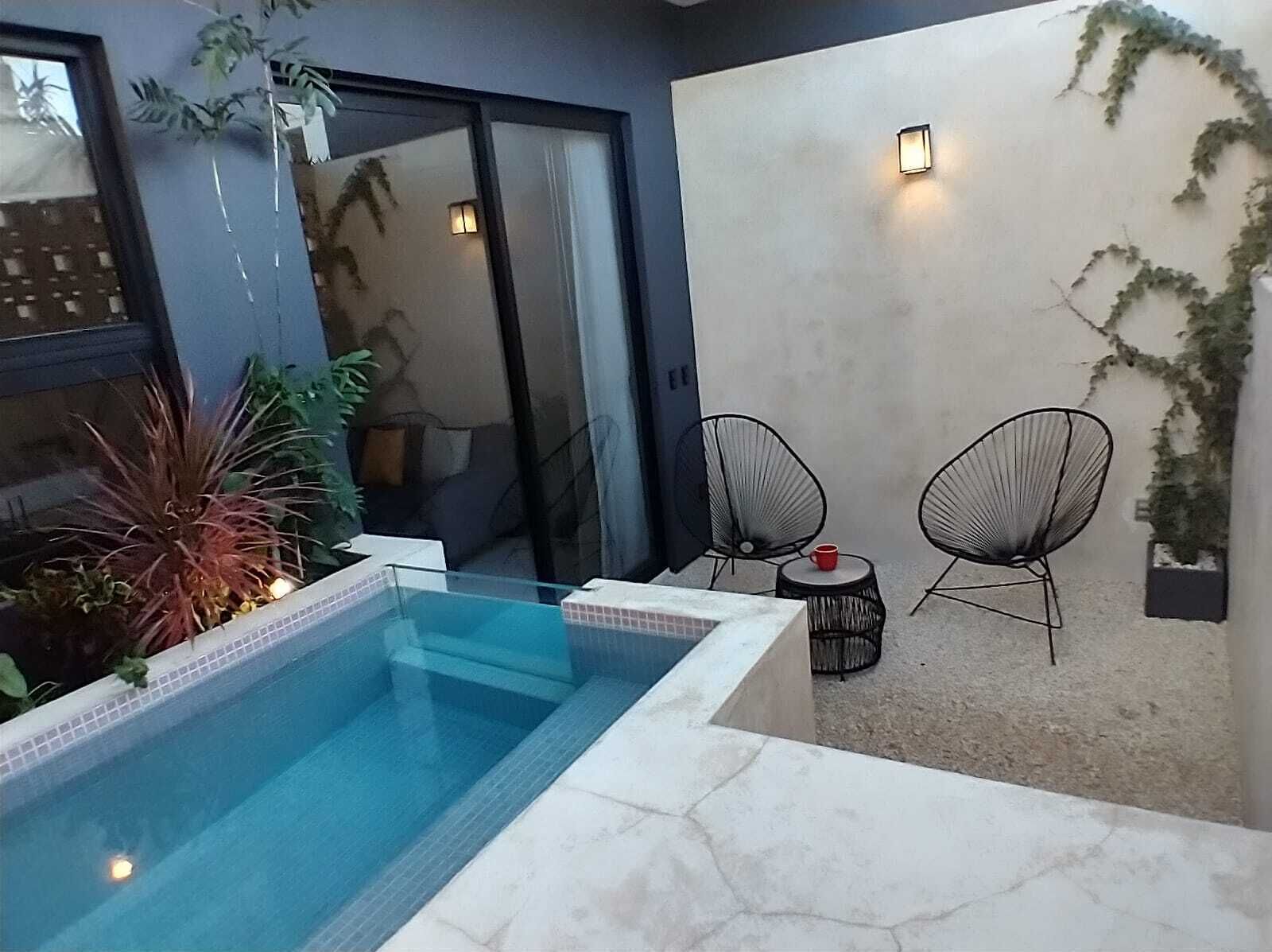 A modern outdoor pool area with a glass enclosure, two black wire chairs, and a small table with a red cup.