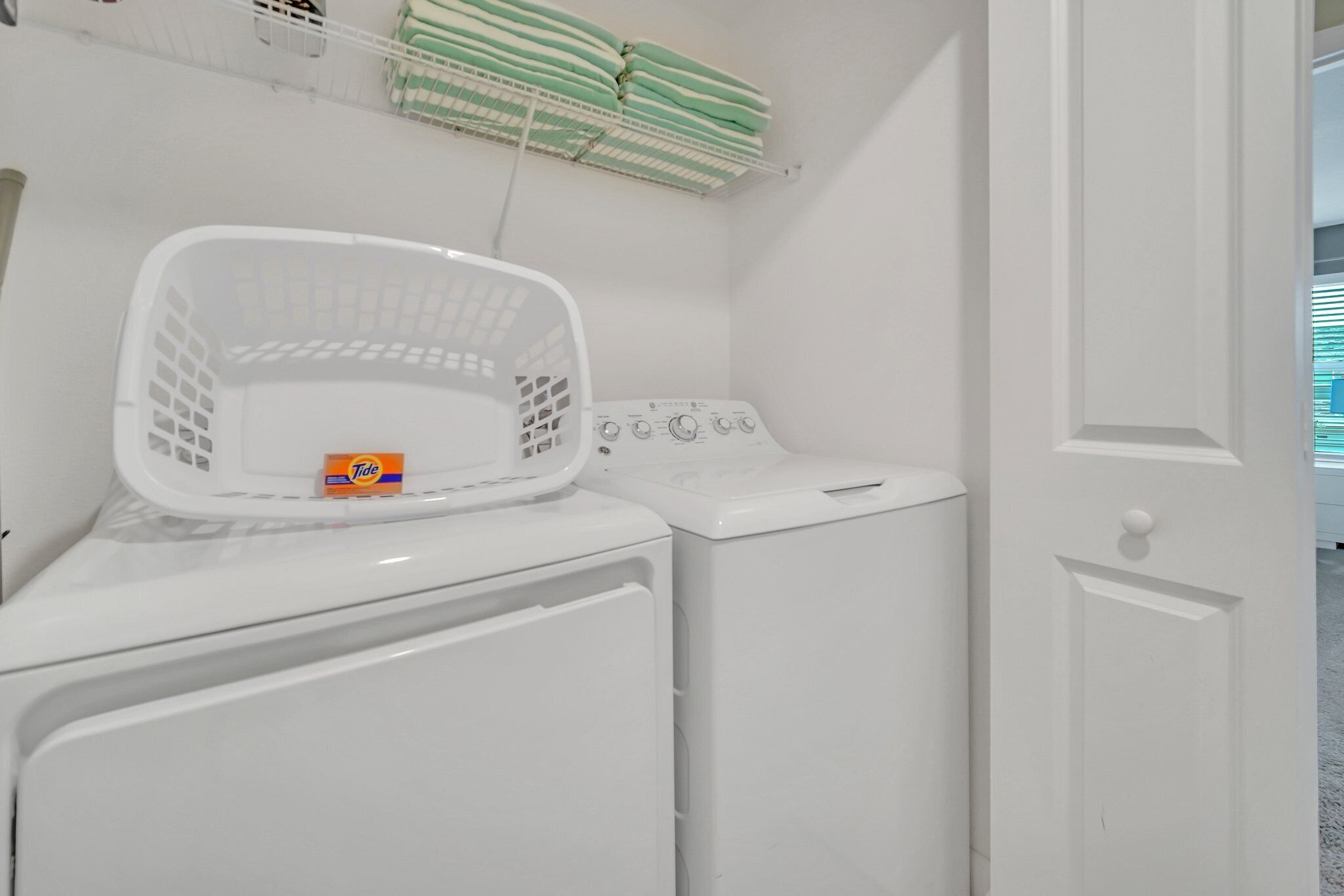 A laundry area with a front-loading washer and dryer, a white basket, and stacked laundry on a shelf.