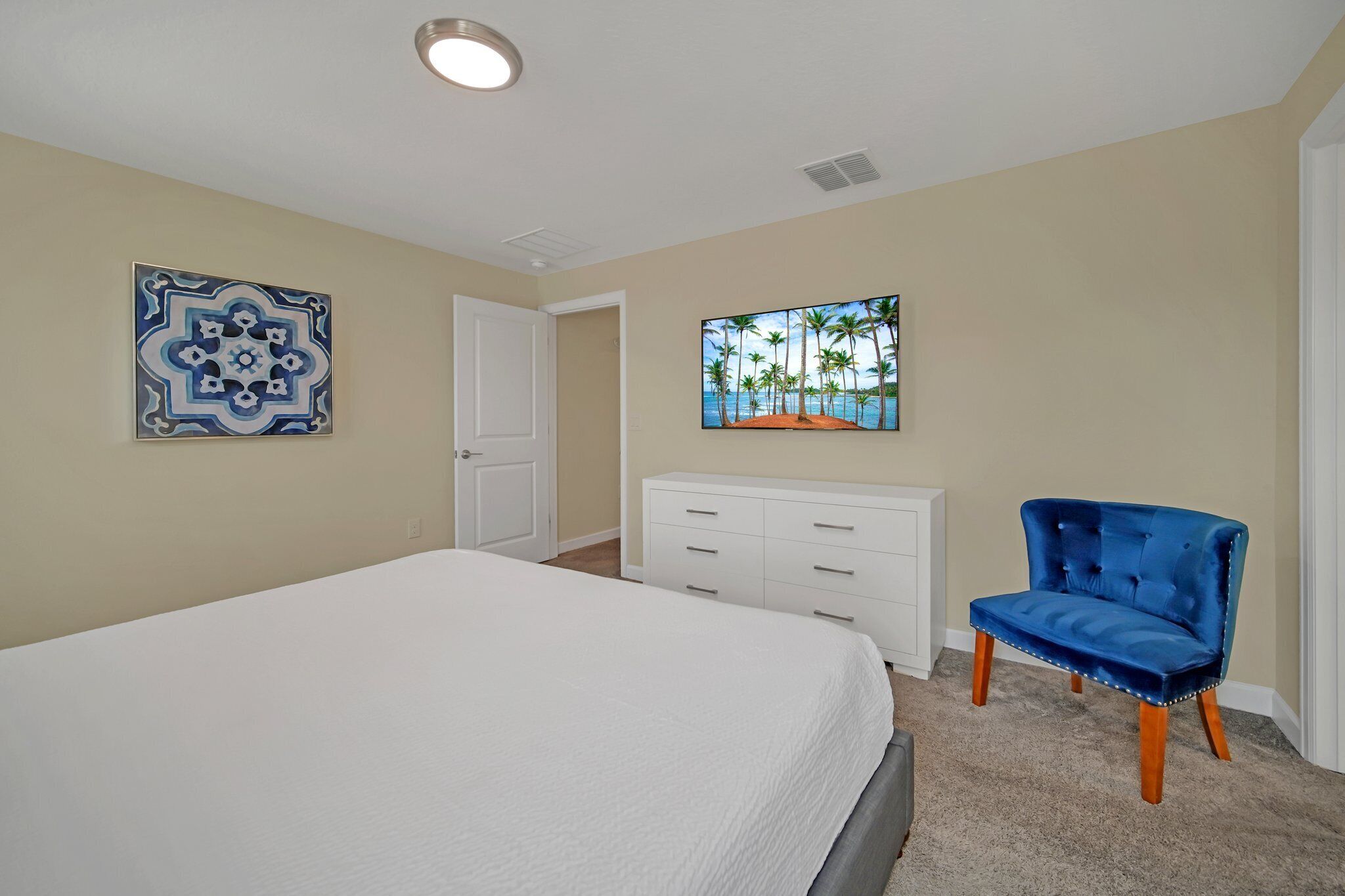 A bedroom with a bed, a white dresser, a blue chair, and two wall art pieces.