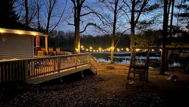 Una terraza de madera con luces colgantes, un espacio cubierto para sentarse y vista a un lago al anochecer.