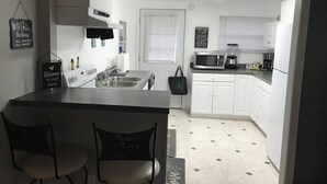 Una cocina con armarios blancos, un microondas, una cafetera y un lavabo. Hay dos sillas y una bolsa negra sobre la encimera.