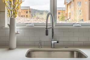 A modern kitchen with a stainless steel sink, a white vase with yellow flowers, and a window with a view of buildings and trees.