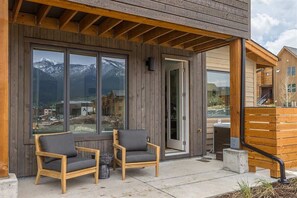 Ein hölzernes Gebäude mit großen Fenstern, die auf Berge blicken, zwei Holzstühle davor und eine Holzterrasse.