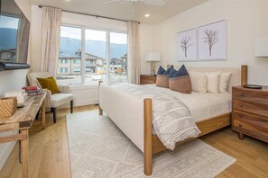 A bedroom with a bed, bedside tables, a desk, and a chair. There is a window with a view of buildings and mountains.