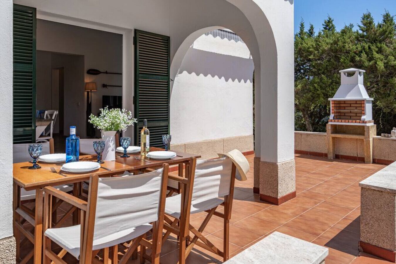 An outdoor patio with a wooden table set for dining, a grill, and a white wall with an archway.