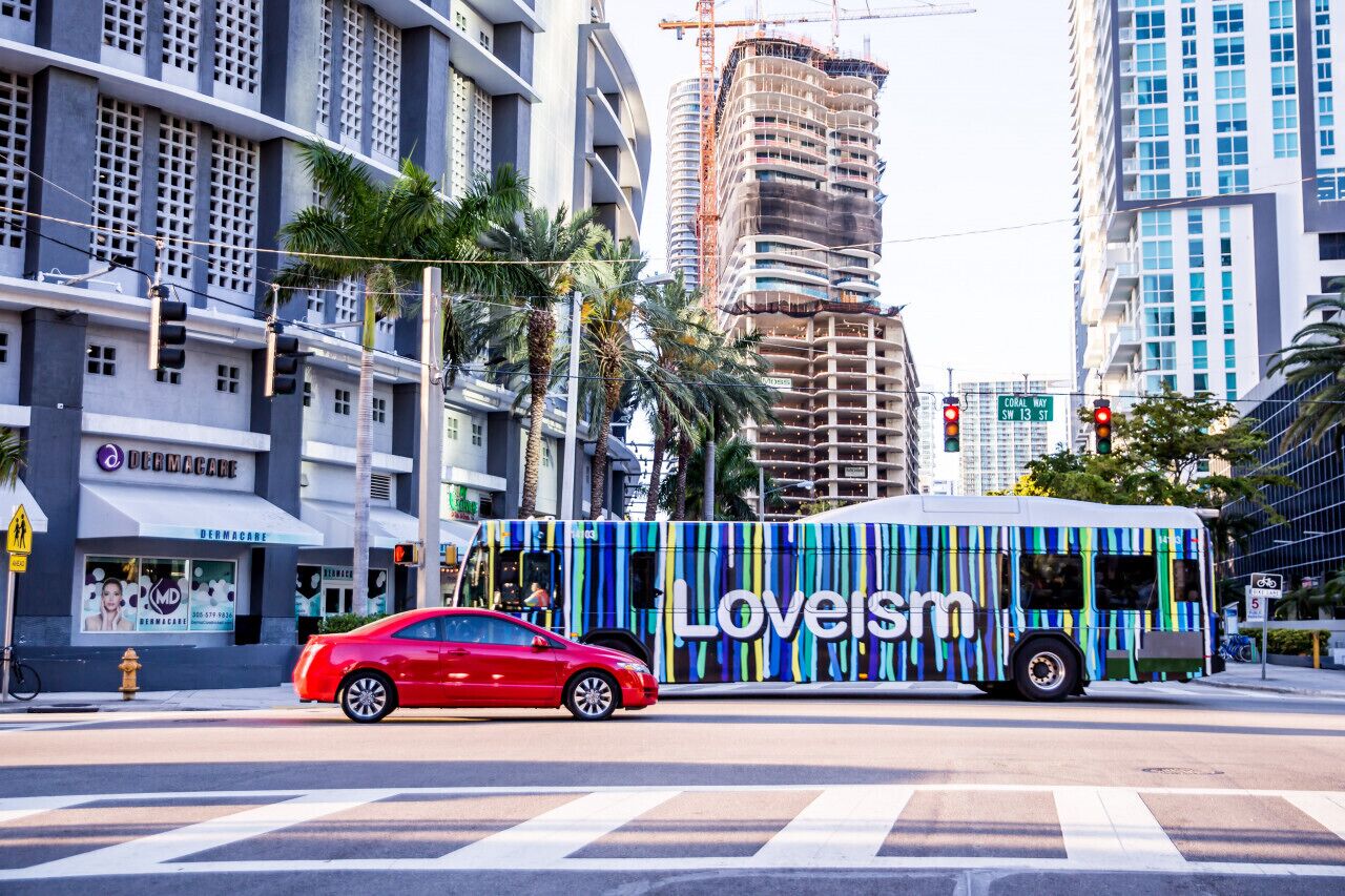 Un auto rojo y un autobús colorido con la leyenda "Loveism" en una intersección urbana con edificios altos y palmeras.