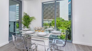 Un comedor moderno con una mesa de vidrio preparada para seis personas, sillas transparentes y un gran ventanal con vistas a edificios y vegetación.