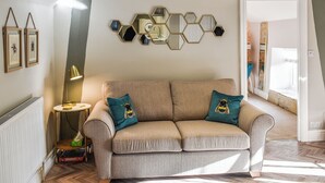 A modern living room with a beige sofa, two teal pillows, a round side table, a floor lamp, and a wall-mounted hexagonal mirror arrangement. - 1 bedroom accommodation in Dronfield (Dronfield)