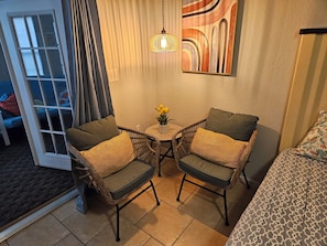 A room with two wicker chairs, a small table with a vase of flowers, a painting on the wall, and a bed with patterned bedding.
