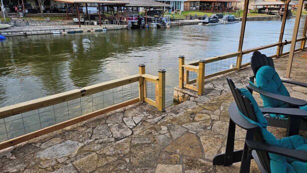 Un patio frente al agua con suelo de piedra, baranda de madera y un área de estar con sillas.