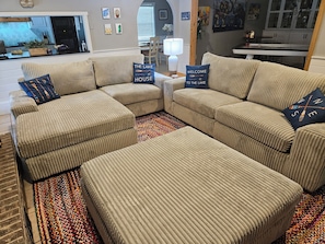 Un salon avec un canapé d’angle beige, deux poufs et un tapis coloré.