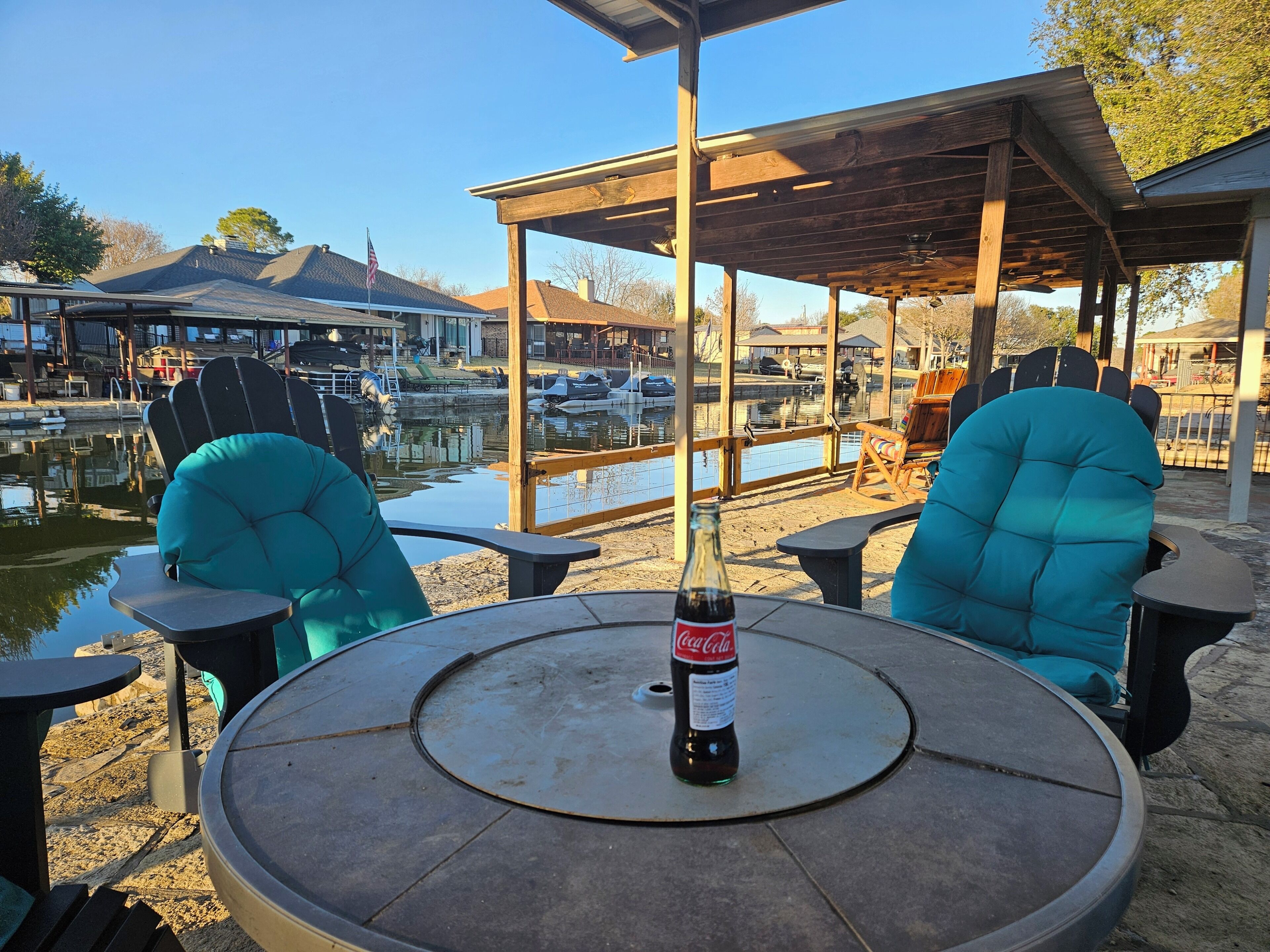 A round table with a bottle of Coca-Cola, two teal chairs, and a wooden pergola structure.
