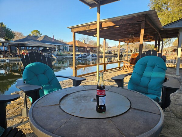 A round table with a bottle of Coca-Cola, two teal chairs, and a wooden pergola structure.