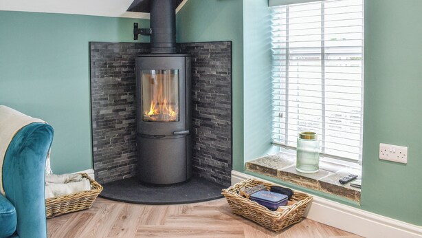 A room with a wood-burning stove, a window with blinds, a blue chair, and a wicker basket on the floor.