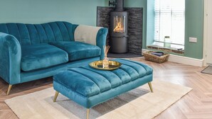 A teal-colored sofa and ottoman set in a living room with a stone fireplace and a window with blinds.