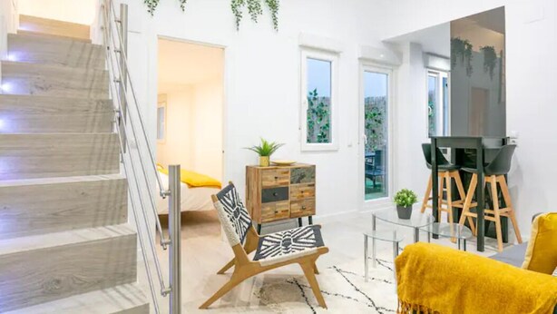 A modern living room with a staircase, a dining area with a black table and chairs, and a large window with a view of greenery. - Your home in the center of Madrid (Madrid)