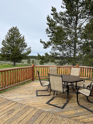 A wooden deck with a table and chairs, surrounded by a wooden railing, overlooking a grassy area with trees.