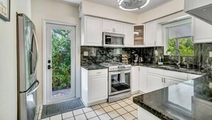 Una cocina moderna con armarios blancos, encimera de mármol negro, electrodomésticos de acero inoxidable y un amplio ventanal que proporciona luz natural.
