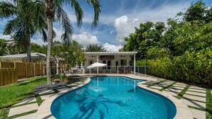 Una piscina con borde curvo, rodeada por una terraza pavimentada con diseños geométricos. Hay un área de patio techada con mobiliario exterior y una pérgola.