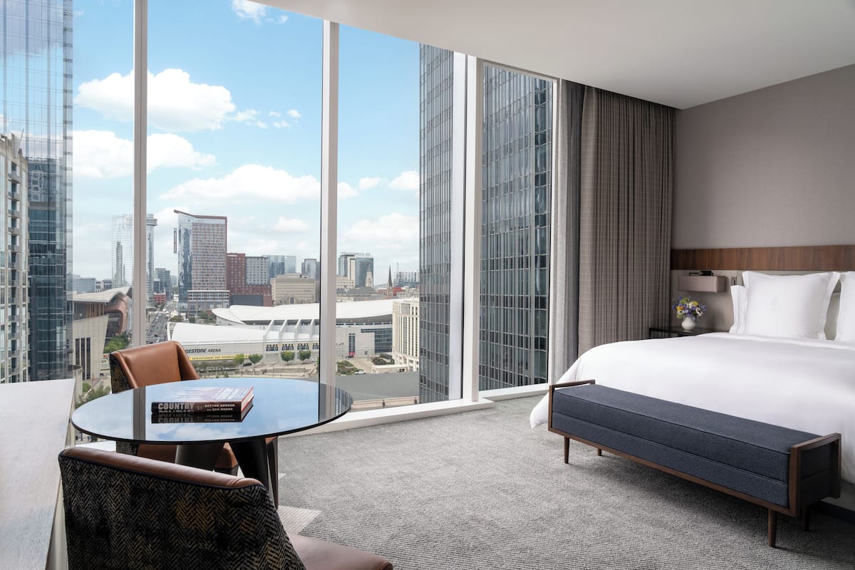A hotel room with a large window, a bed, a bedside table, a chair, and a small table with a book.