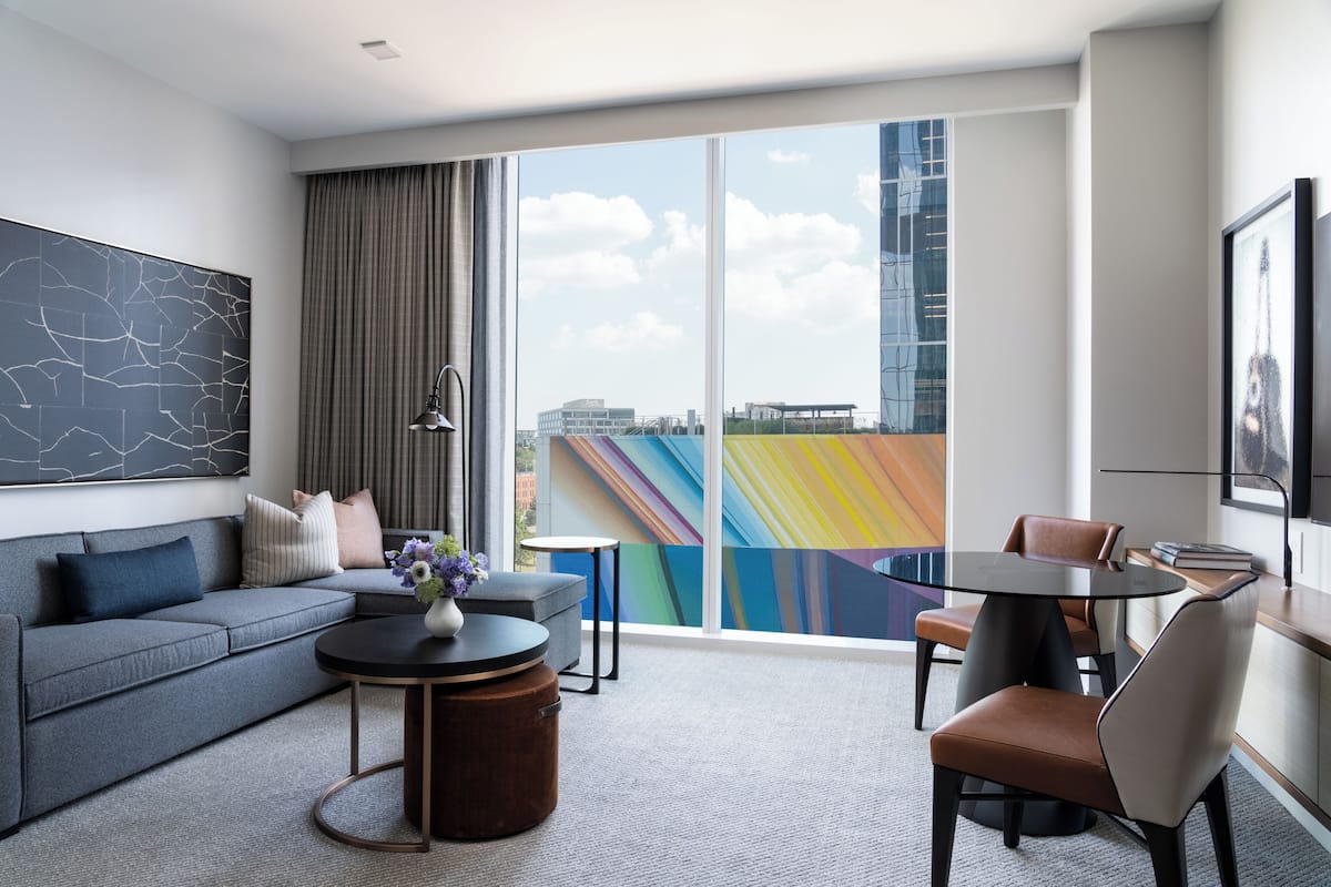 A modern hotel room with a grey sofa, a round coffee table, and a dining area with a table and chairs.