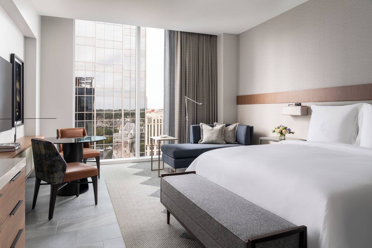 A modern hotel room with a large bed, a desk with a chair, a small table, and a view of the city through large windows.
