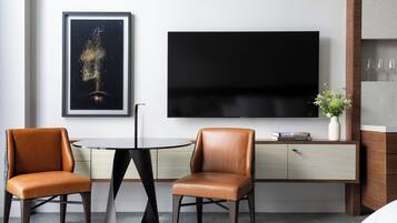 A modern room with a round table, two leather chairs, a flat-screen TV, a framed artwork, a cabinet with wine glasses, and a vase with flowers.