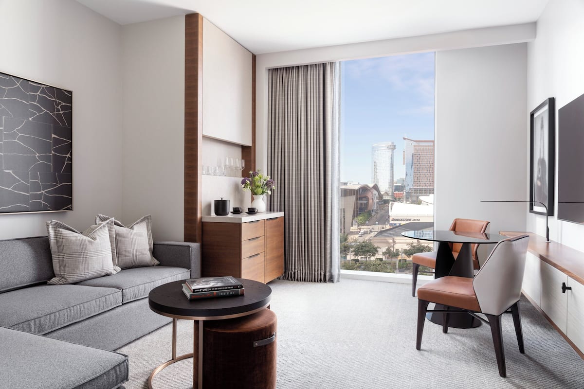A modern hotel room with a grey sofa, a round coffee table, a dining area with a chair, a wooden cabinet, and a large window offering a city view.