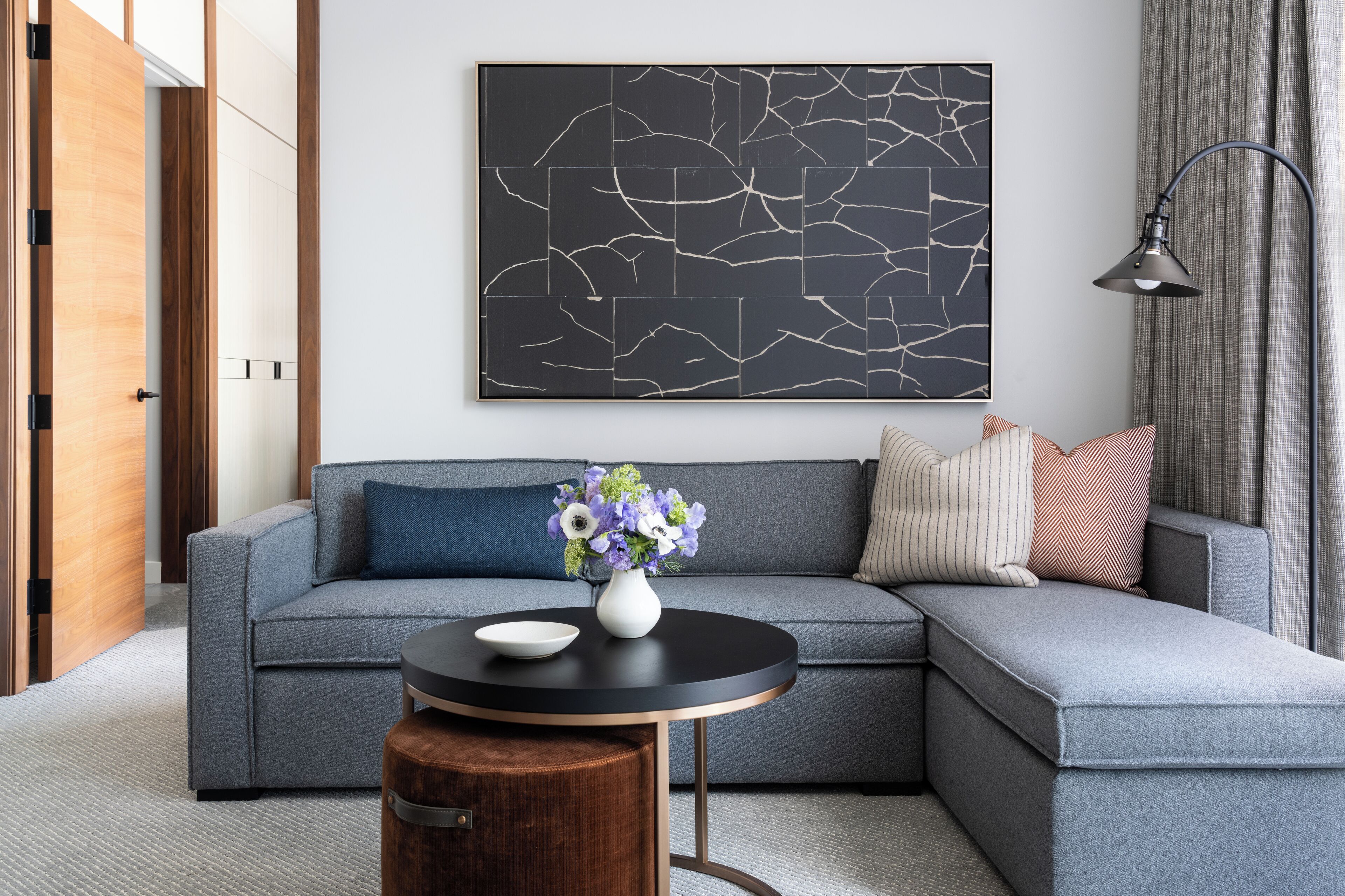 A modern living room with a grey sectional sofa, a round wooden coffee table, and a black abstract painting on the wall.