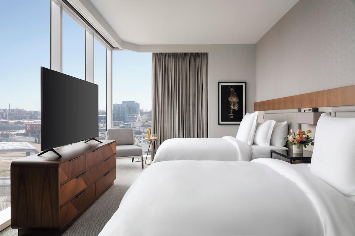 A hotel room with a large TV on a wooden dresser, two beds, a chair, a small table with a vase of flowers, and a city view through large windows.