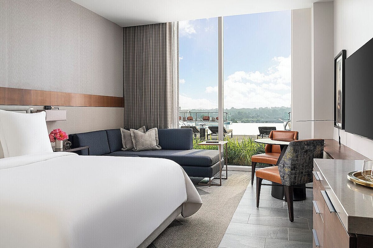 A modern hotel room with a large bed, a sofa, a dining area, and a balcony with a view.