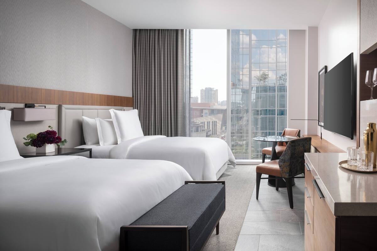 A modern hotel room with two beds, a dining area, and a large window offering a city view.