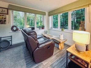 A modern living room with a leather recliner, a wooden coffee table, and a lamp. Large windows provide a view of the greenery outside.