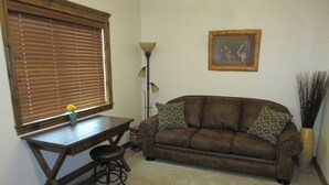 A living room with a brown leather sofa, a wooden coffee table, a small stool, a floor lamp, a framed picture on the wall, and a vase with a broom. - Dual Queens Townhomes On the Green (Afton)