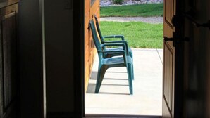 An open doorway leading to a wooden exterior with a stone wall and a grassy area with two green plastic chairs. - Dual Queens Townhomes On the Green (Afton)
