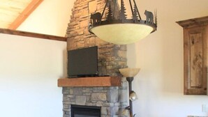 A cozy living room with a stone fireplace, a round wooden table, and a patterned armchair. - Dual Queens Townhomes On the Green (Afton)