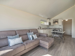 A modern living room with a sectional sofa, a dining table with chairs, a kitchen area with white cabinets, and a ceiling with linear lighting.