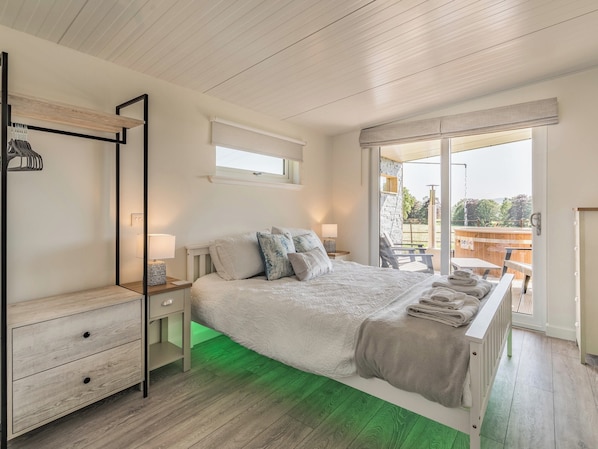 A bedroom with a bed, a bedside table, a hanging rack, and a view of an outdoor deck.