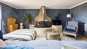 Une chambre confortable avec une cheminée en pierre, un lit recouvert d’une couverture à rayures, une table de chevet en bois et un fauteuil gris.