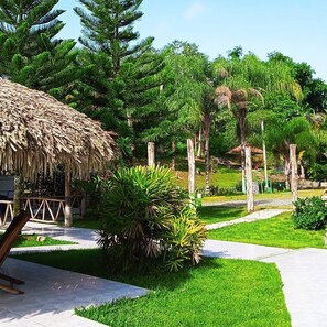 A tropical garden with a thatched-roof structure, palm trees, and a paved walkway. - Serapius Green (Sabana Grande De Boyá)