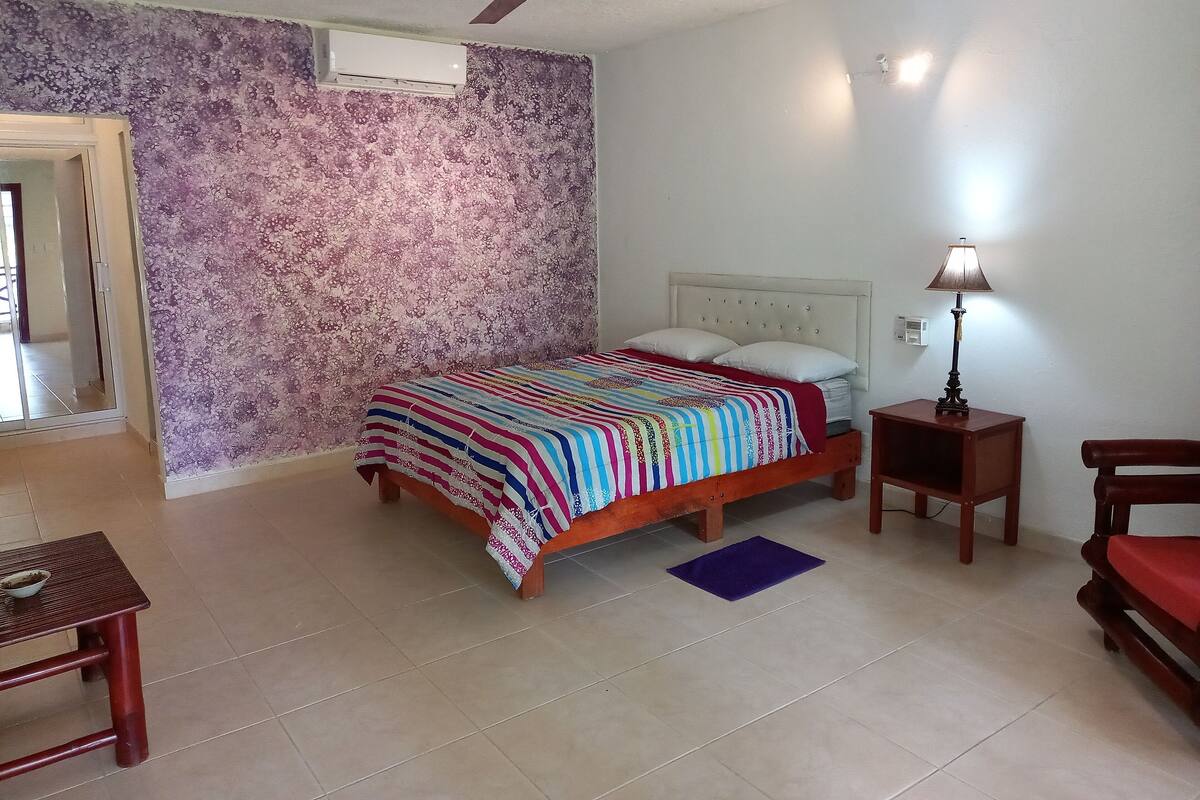 Een slaapkamer met een bed, een plafondventilator, een nachtkastje, een lamp en een stoel.